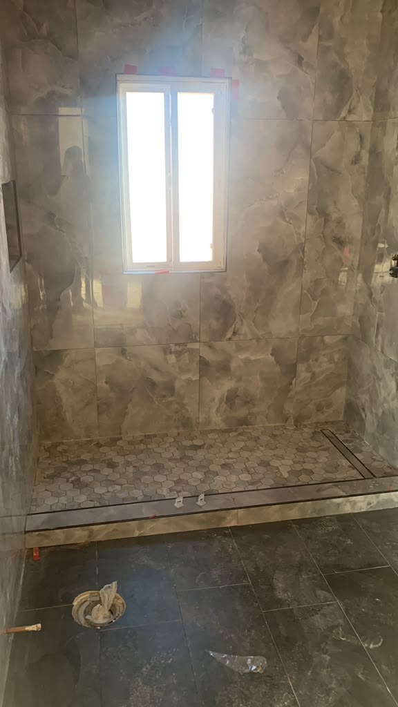 A modern bathroom with gray marble tiles, a window, and a glass shower enclosure. The floor features hexagonal tiles.