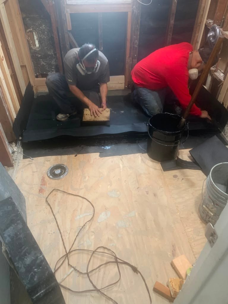 Two workers in a partially renovated bathroom applying waterproofing material on the floor, surrounded by construction tools and materials.