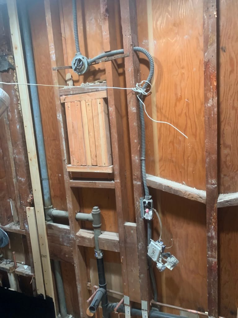 Exposed bathroom wall showing wooden framing, electrical wiring, and plumbing pipes, indicating a remodeling project in progress.