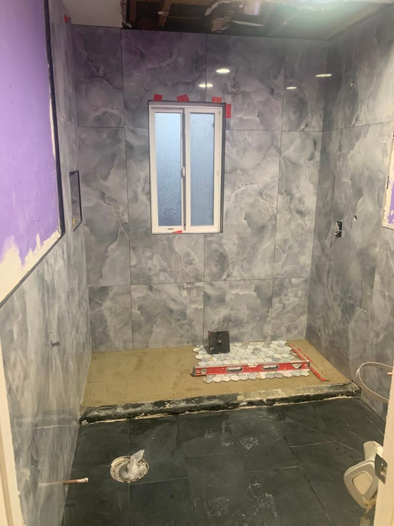 A partially remodeled bathroom featuring gray marble walls, a window, and an unfinished floor with tiles and tools visible.