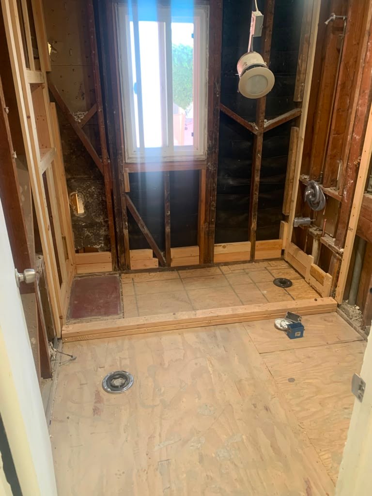 Bathroom under renovation, featuring exposed framing, a window, and unfinished flooring with plumbing fixtures visible.