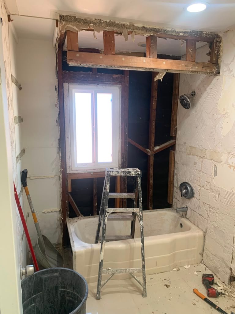 A partially renovated bathroom showing exposed framing, a bathtub, a ladder, and tools, with a window in the background.