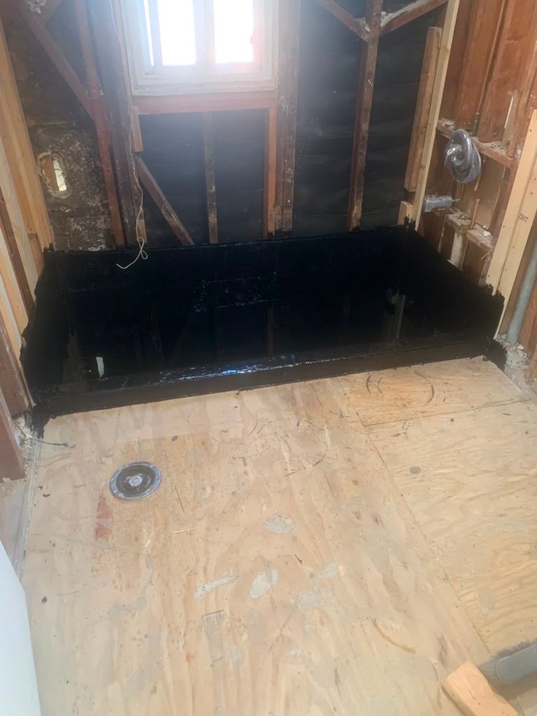 A partially renovated bathroom showing a black waterproofed shower base on a plywood floor, with exposed framing and plumbing.