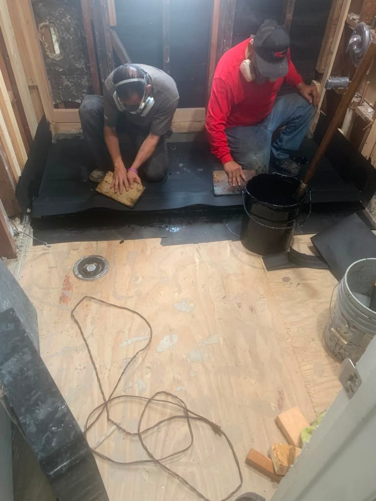 Two workers in a partially renovated bathroom, applying materials to the floor. Tools and construction materials are visible.