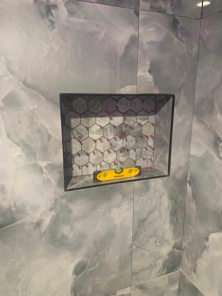 A shower niche with hexagonal tiles and a yellow level tool, set against a marbled gray wall.