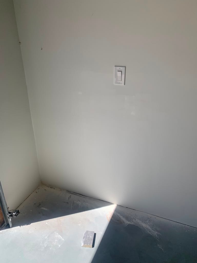 A corner of a garage with a light switch on the wall, showing a dry, unfinished floor and sunlight streaming in from an open door.