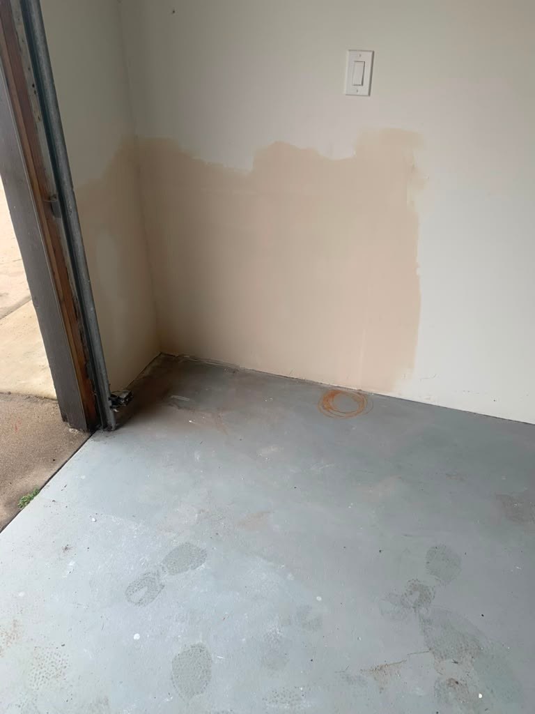 A corner of a garage with a partially painted wall showing water damage, a concrete floor with visible footprints, and a light switch.