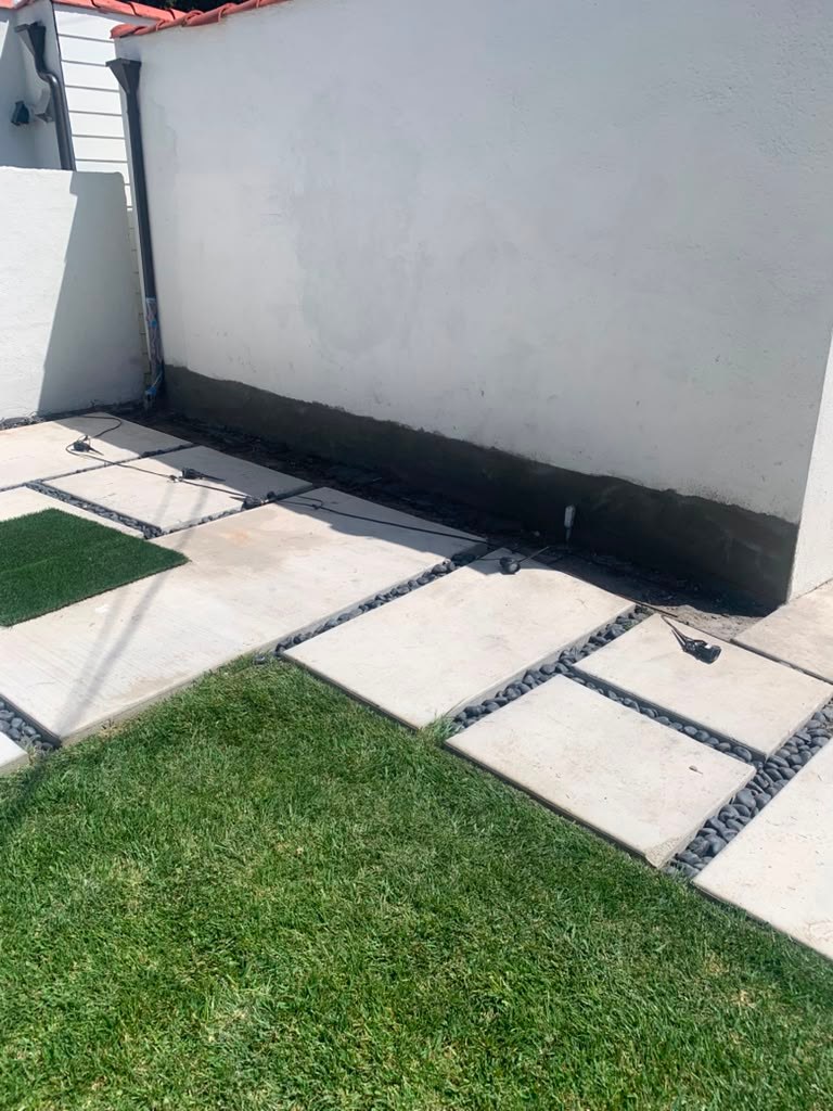 A white garage wall with visible water damage, adjacent to a paved area and green grass, showing potential waterproofing issues.