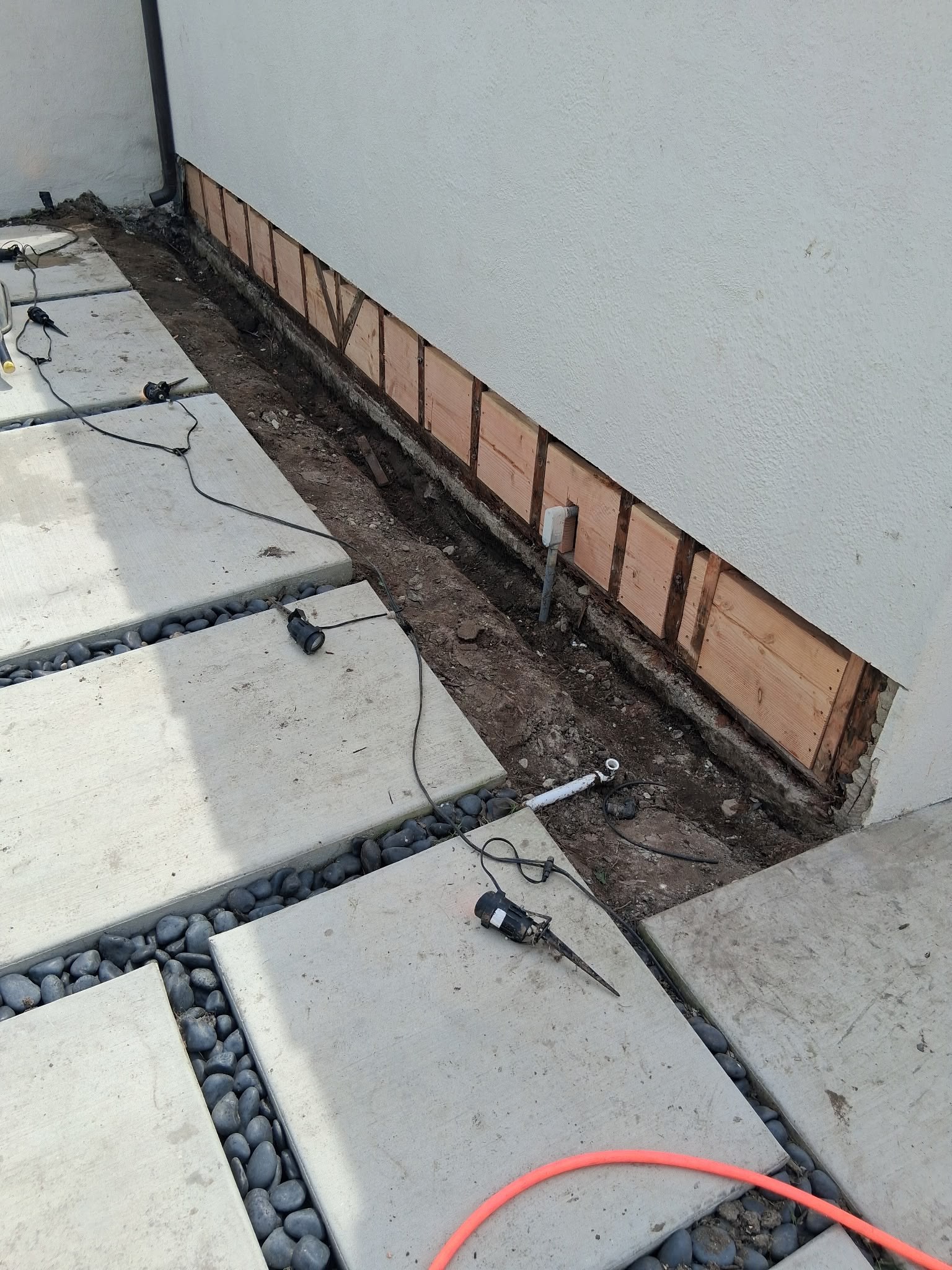 Leaking garage wall with exposed wooden framing, gravel, and concrete pavers, showing tools and equipment nearby.