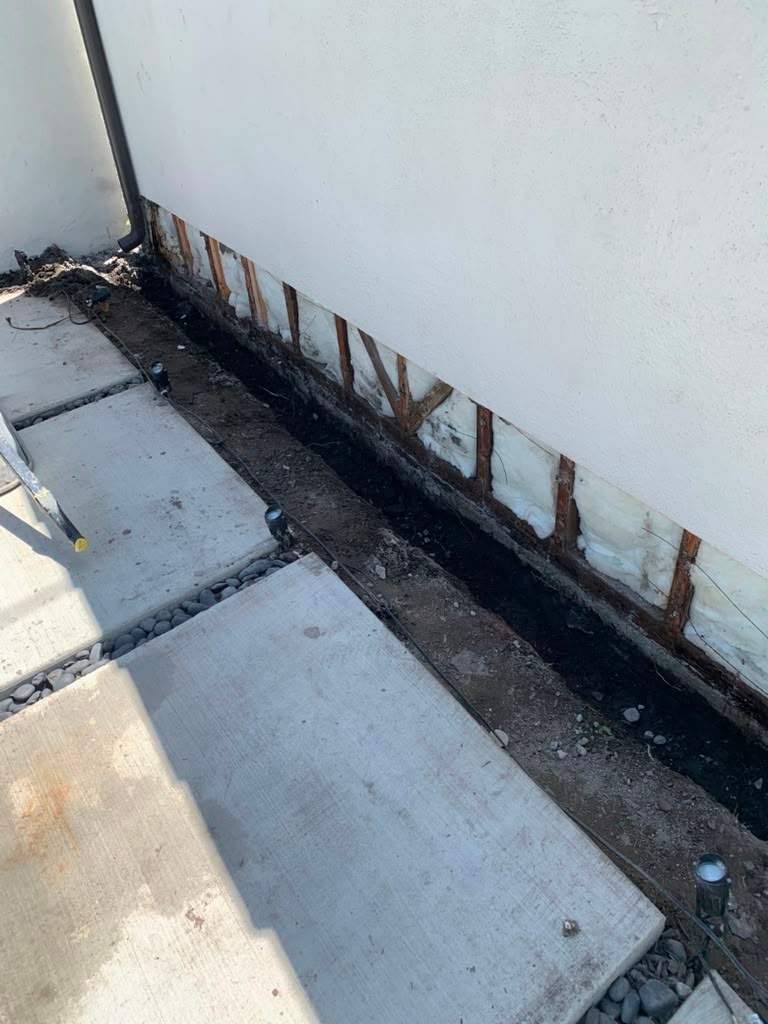 A section of an exterior garage wall shows exposed framing and a trench with gravel, indicating water damage and repair work.