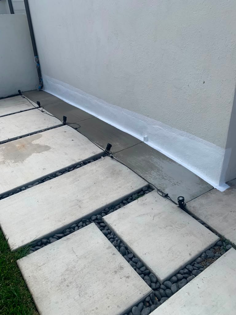 A garage wall with visible water damage, featuring a white waterproofing membrane along the base, surrounded by stone and concrete pavers.