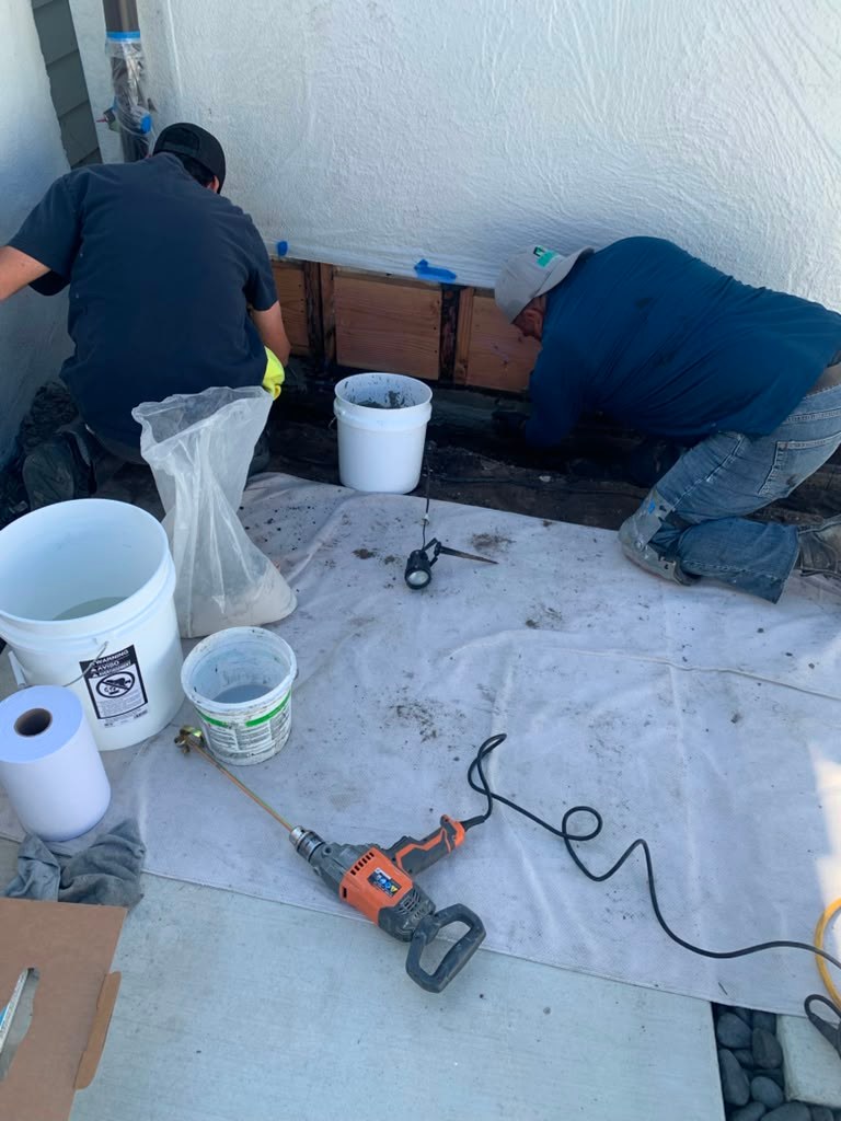 Two workers kneel beside a garage wall, repairing a leak. Tools, buckets, and a tarp are visible on the ground.