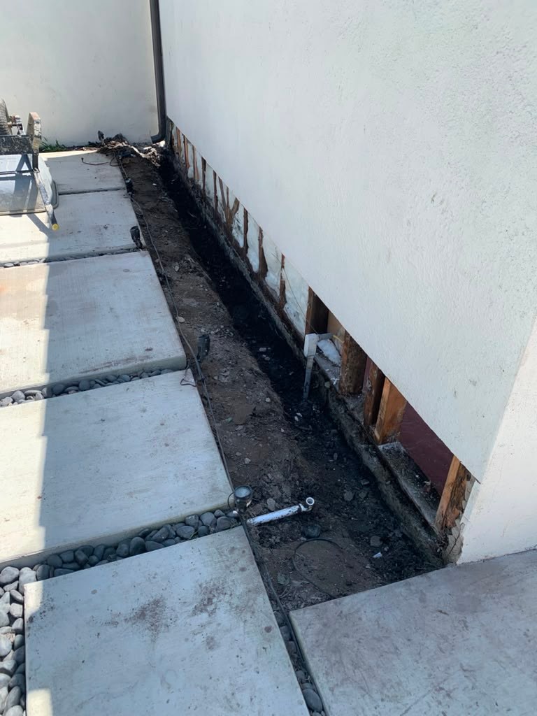 A partially exposed garage wall shows water damage with exposed wood framing and a gravel pathway beside it.