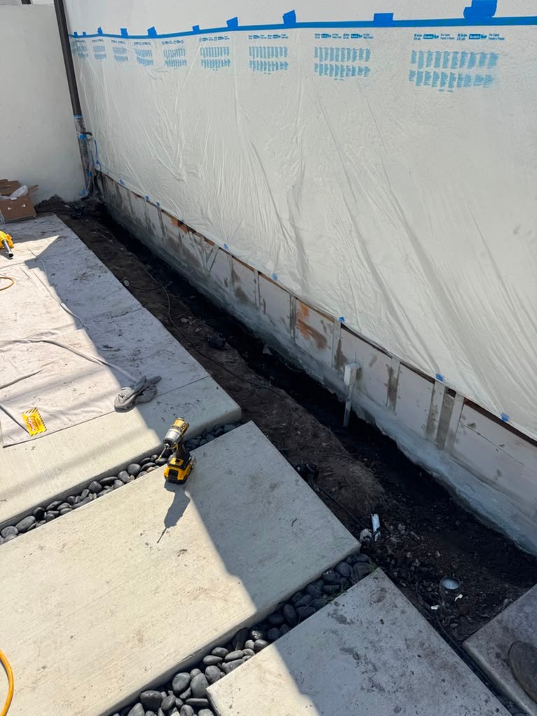 A garage wall with visible water damage, exposed foundation, and construction materials, including tools and a gravel path.