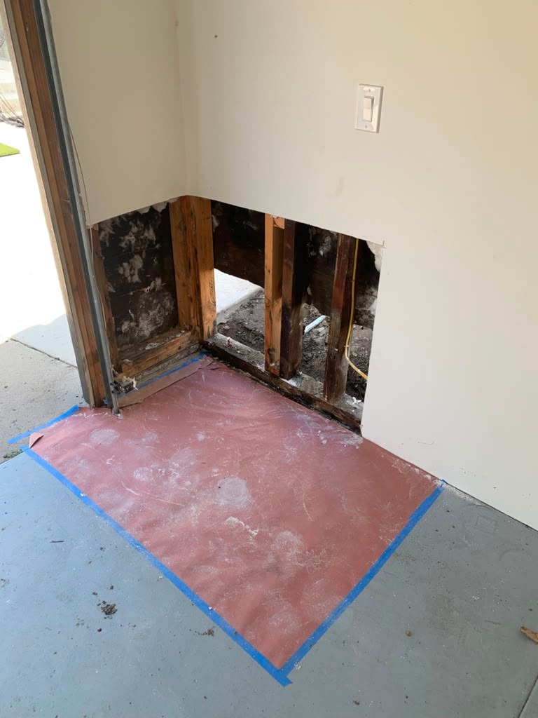 A corner of a garage with exposed wall framing, showing water damage and mold, and a protective mat on the floor.