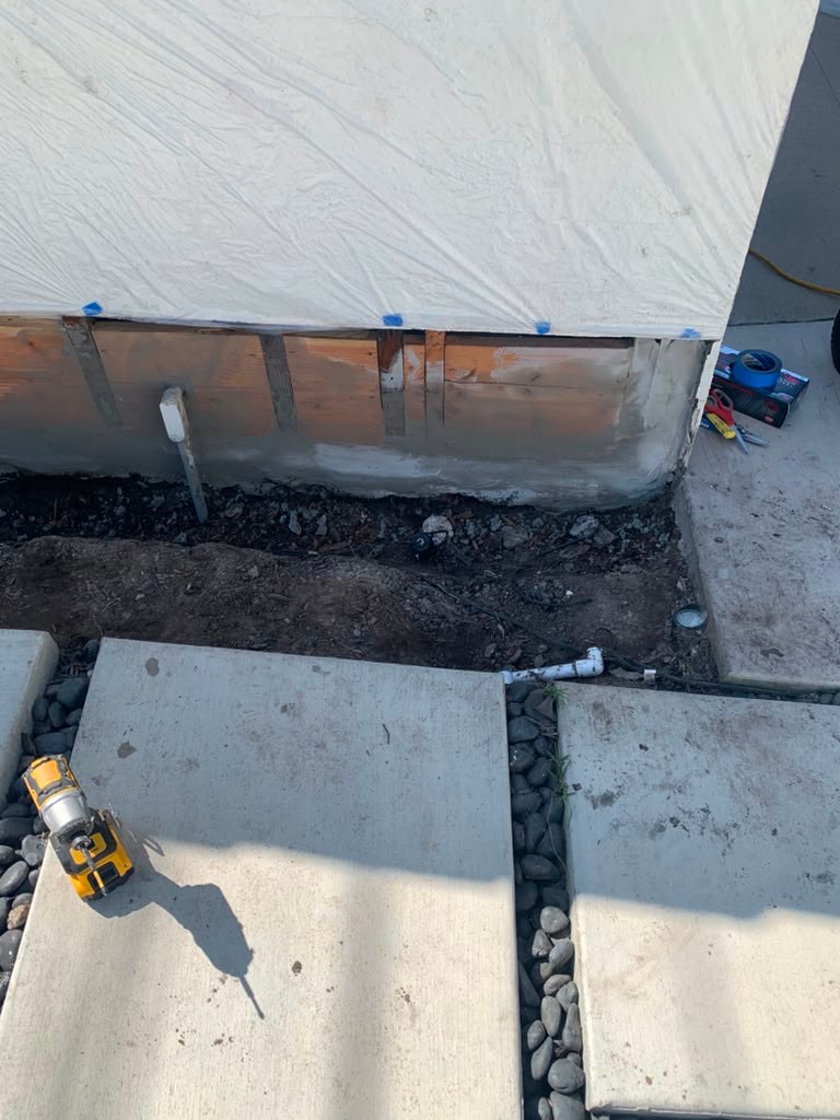 Exterior garage wall with visible leaks, exposed foundation, and a partially covered area. Concrete pathway and tools nearby.