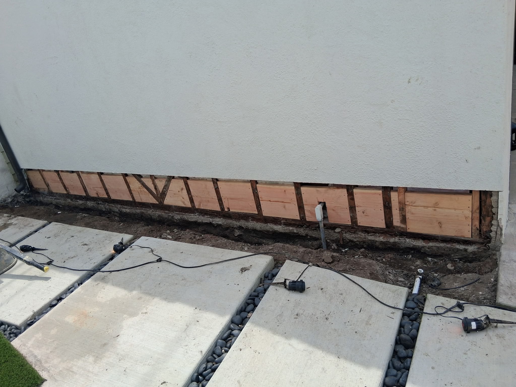 A garage wall with exposed wooden framing and a trench below, surrounded by concrete slabs and electrical wires.