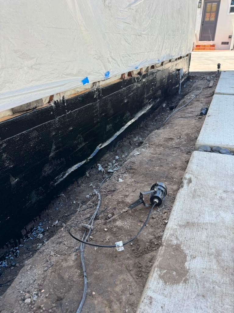 A partially excavated garage wall with visible black waterproofing material, surrounded by dirt and concrete, showing signs of water damage.
