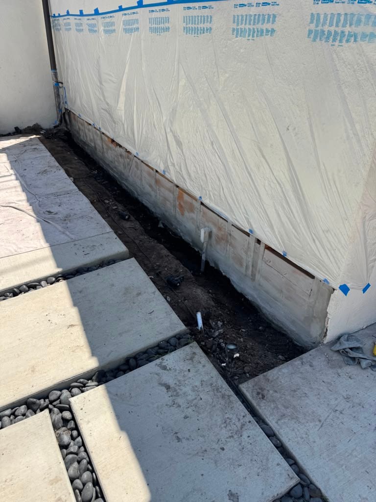 A garage wall with a plastic covering shows a gap at the base, revealing dirt and gravel, indicating potential water damage.