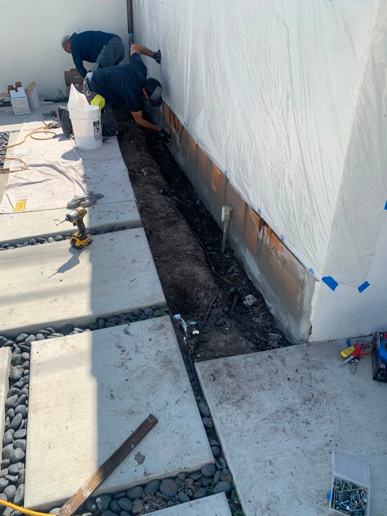 Two workers are repairing a leaking garage wall, digging a trench along the base of a white wall, with tools and materials nearby.