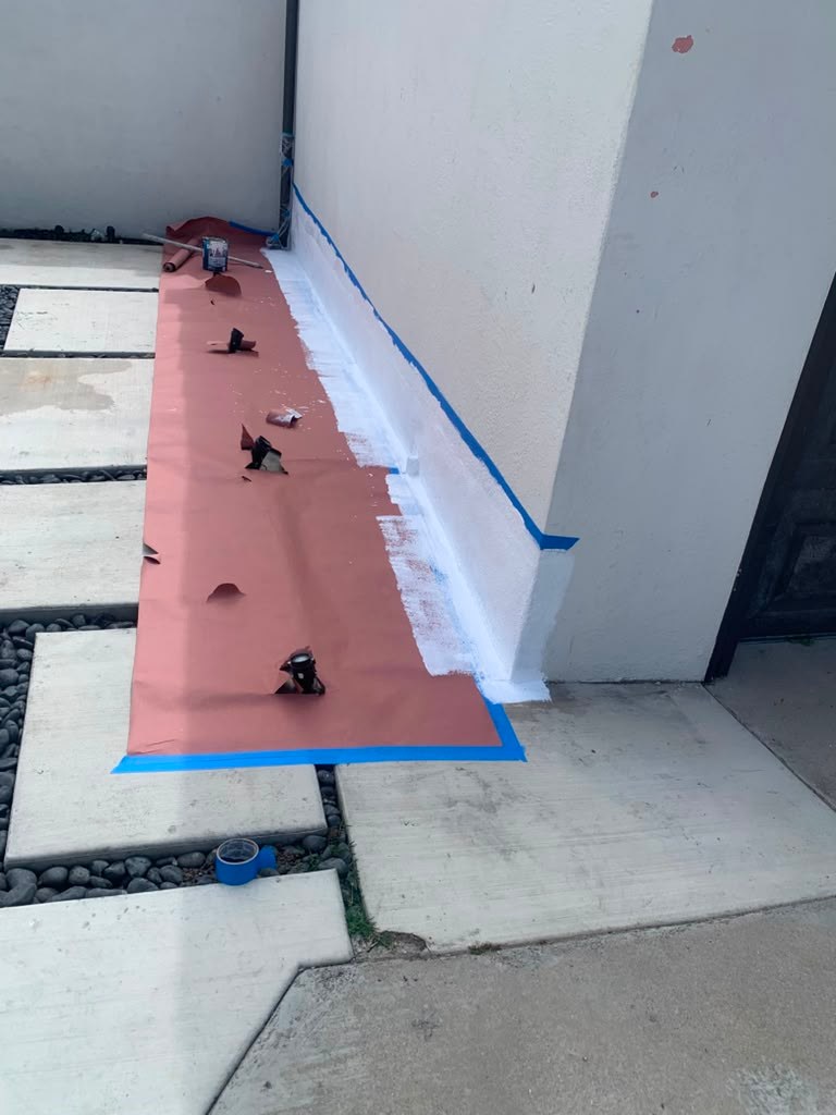 Exterior garage wall with a waterproofing membrane applied, blue tape along the edge, and a concrete pathway with pebbles.