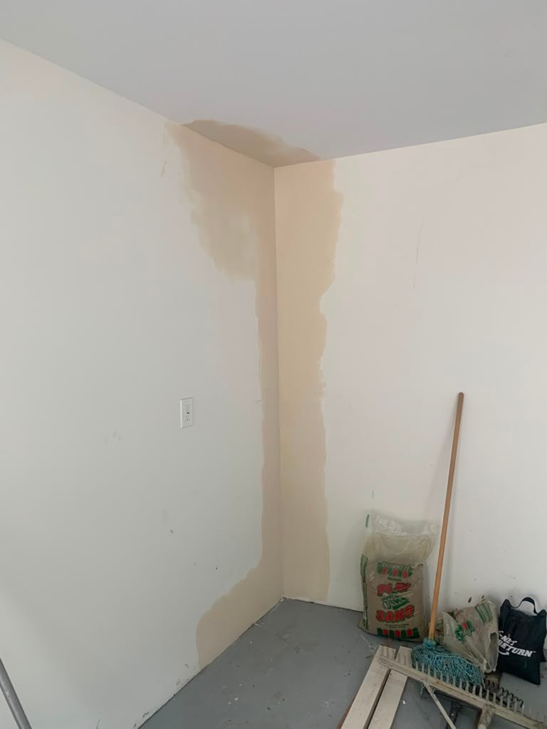 A corner of a garage wall shows water stains and discoloration, with a broom and bag visible on the floor.
