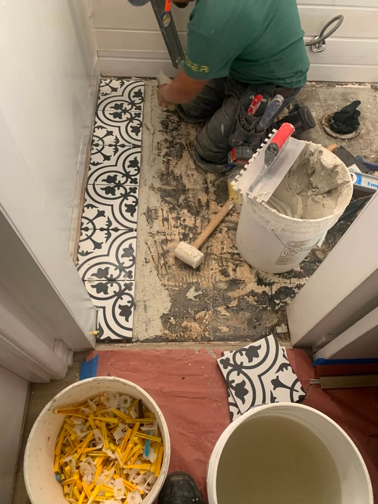 A contractor repairs an uneven bathroom tile floor, using tools and materials like a mallet and buckets of grout on a patterned tile surface.