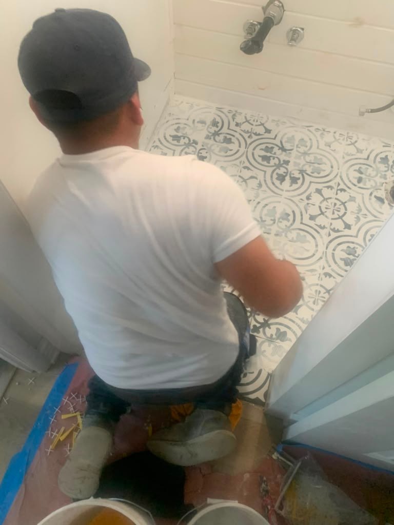 A person kneels on a tiled bathroom floor, working on repairs. Tools and buckets are visible nearby, with patterned tiles in the background.