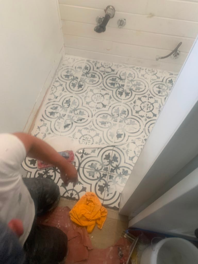 A person kneels on a patterned tile bathroom floor, applying grout. Tools and cleaning cloths are scattered nearby.