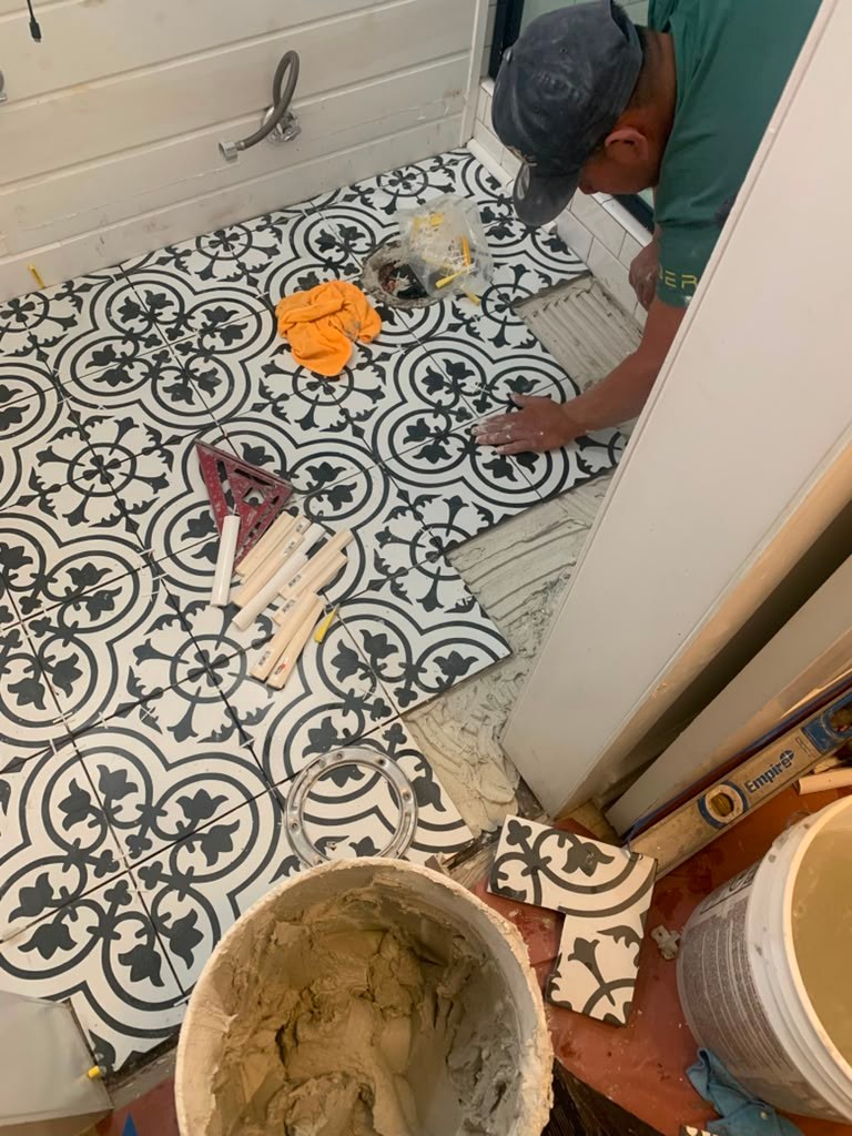 A person installs patterned black and white tiles on a bathroom floor, with tools and materials scattered around, including a bucket of mortar.