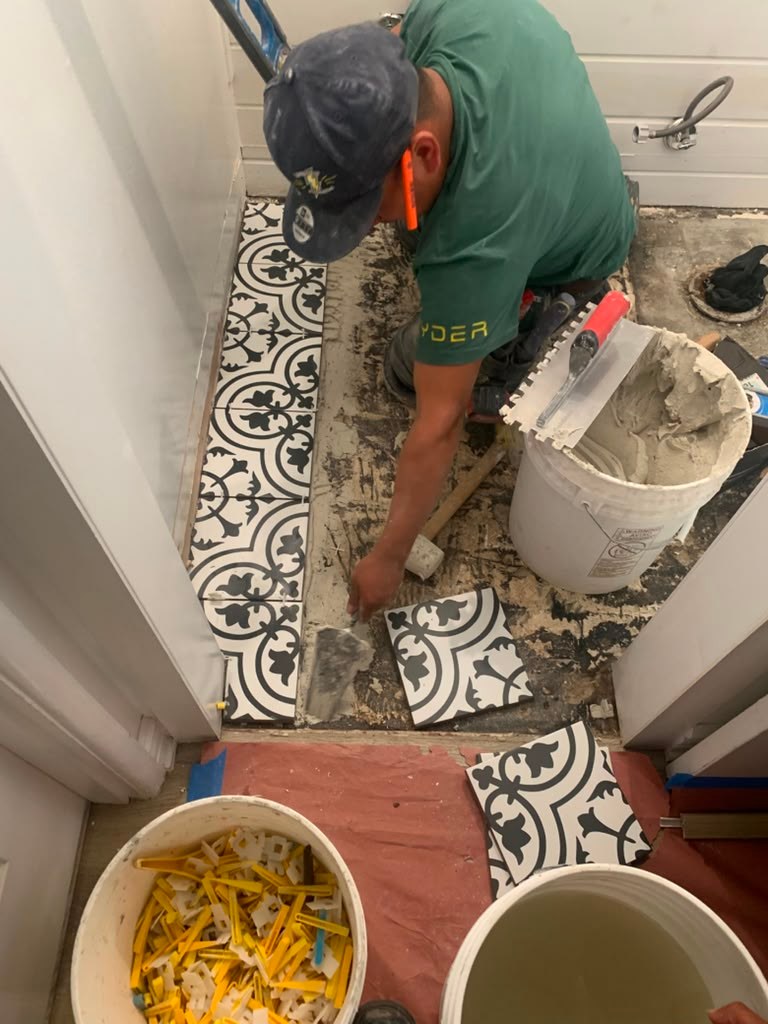 A worker in a green shirt lays patterned tiles on a bathroom floor, with a bucket of mortar and tools nearby.