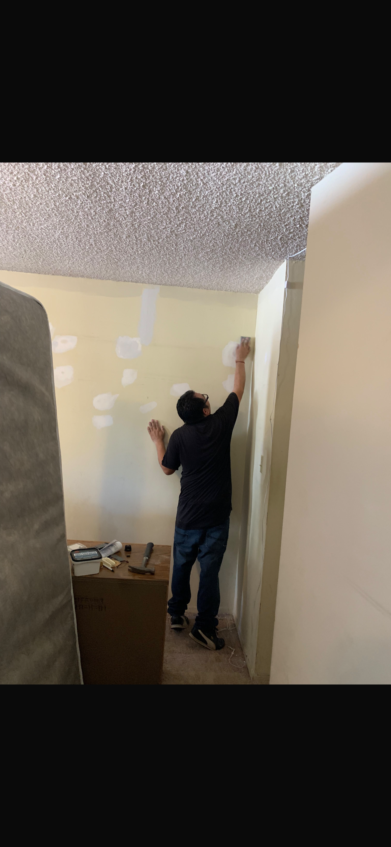 A person in a black shirt applies joint compound to a wall in a room with unfinished drywall and tools on a nearby surface.
