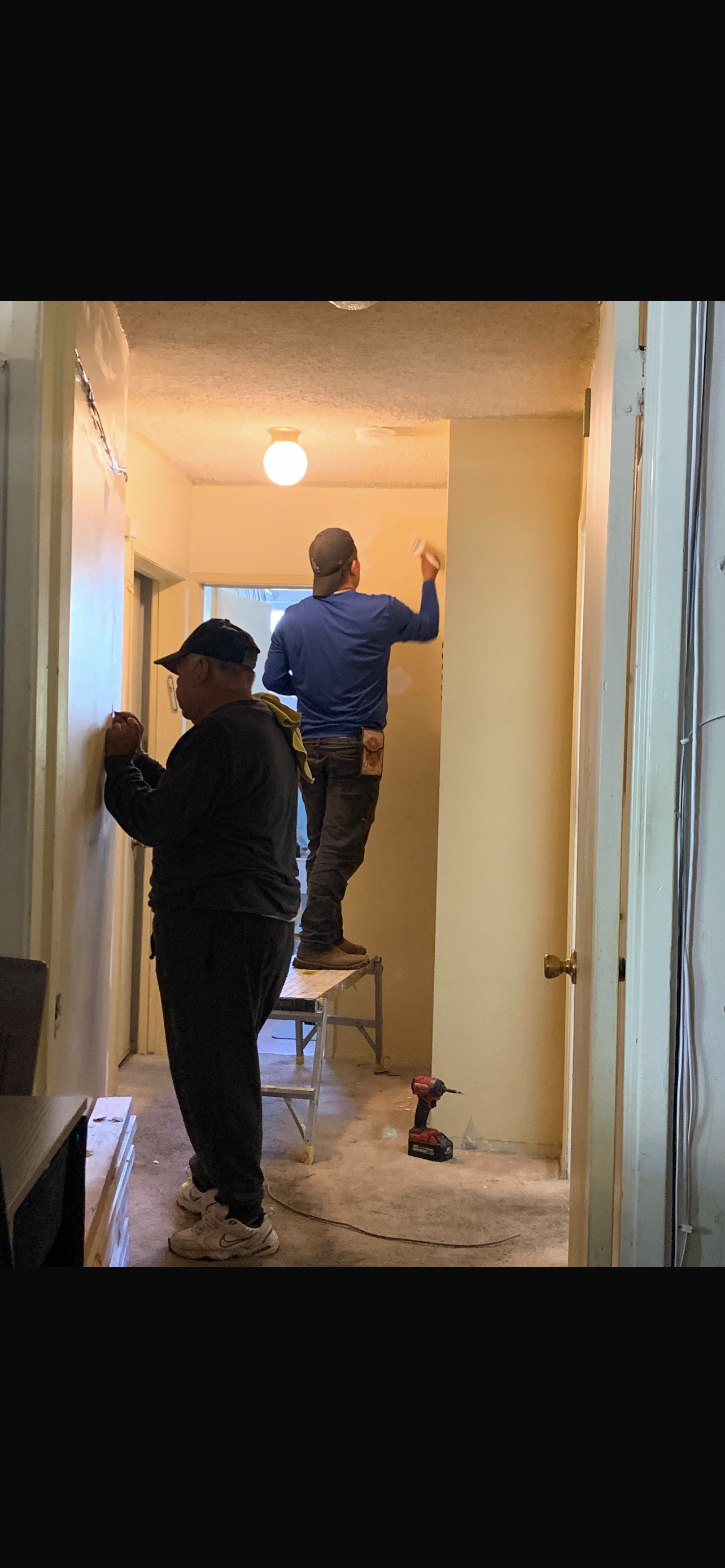 Two men working in a hallway; one stands on a stool painting a wall, while the other uses a tool on a lower wall.