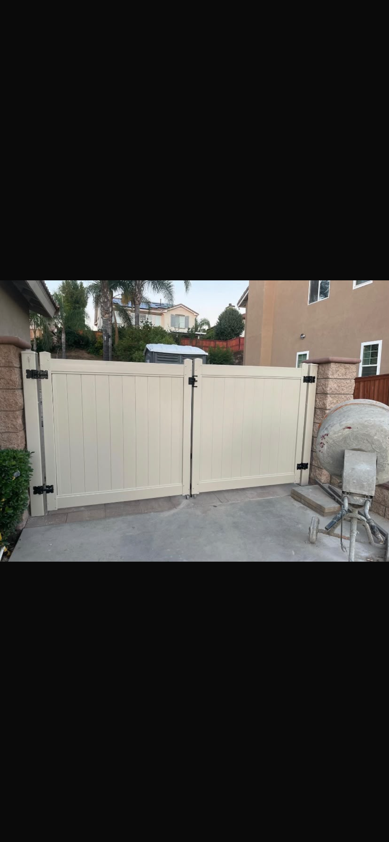 A vinyl double gate with a light beige color, flanked by a stone wall, set in a residential area with nearby houses and greenery.