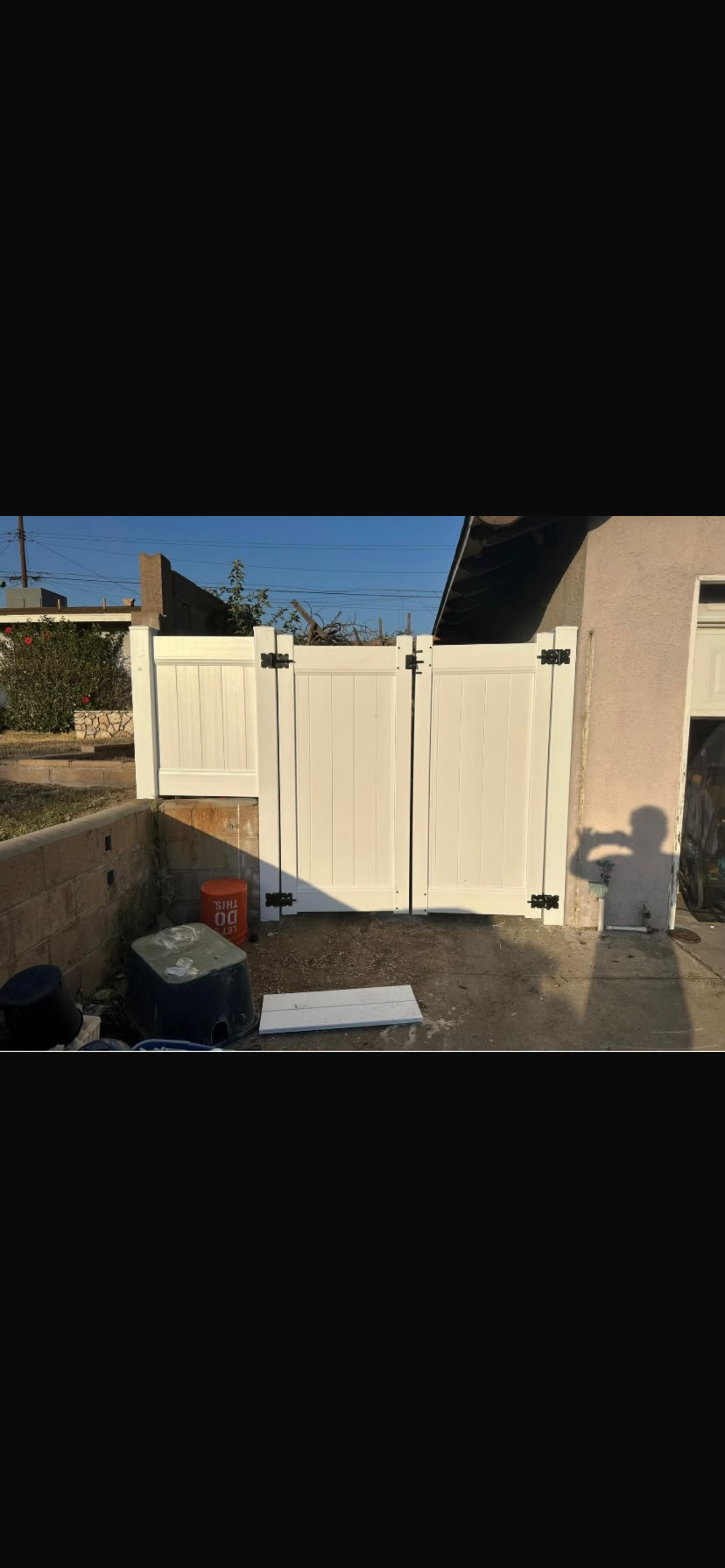 A white vinyl double gate is installed in a residential setting, with a concrete surface and a clear blue sky in the background.
