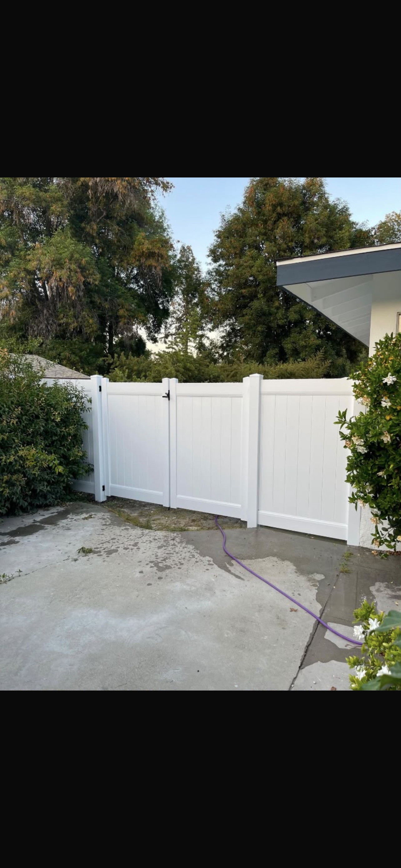A white vinyl fence with a double gate encloses a concrete area, surrounded by greenery and trees in the background.
