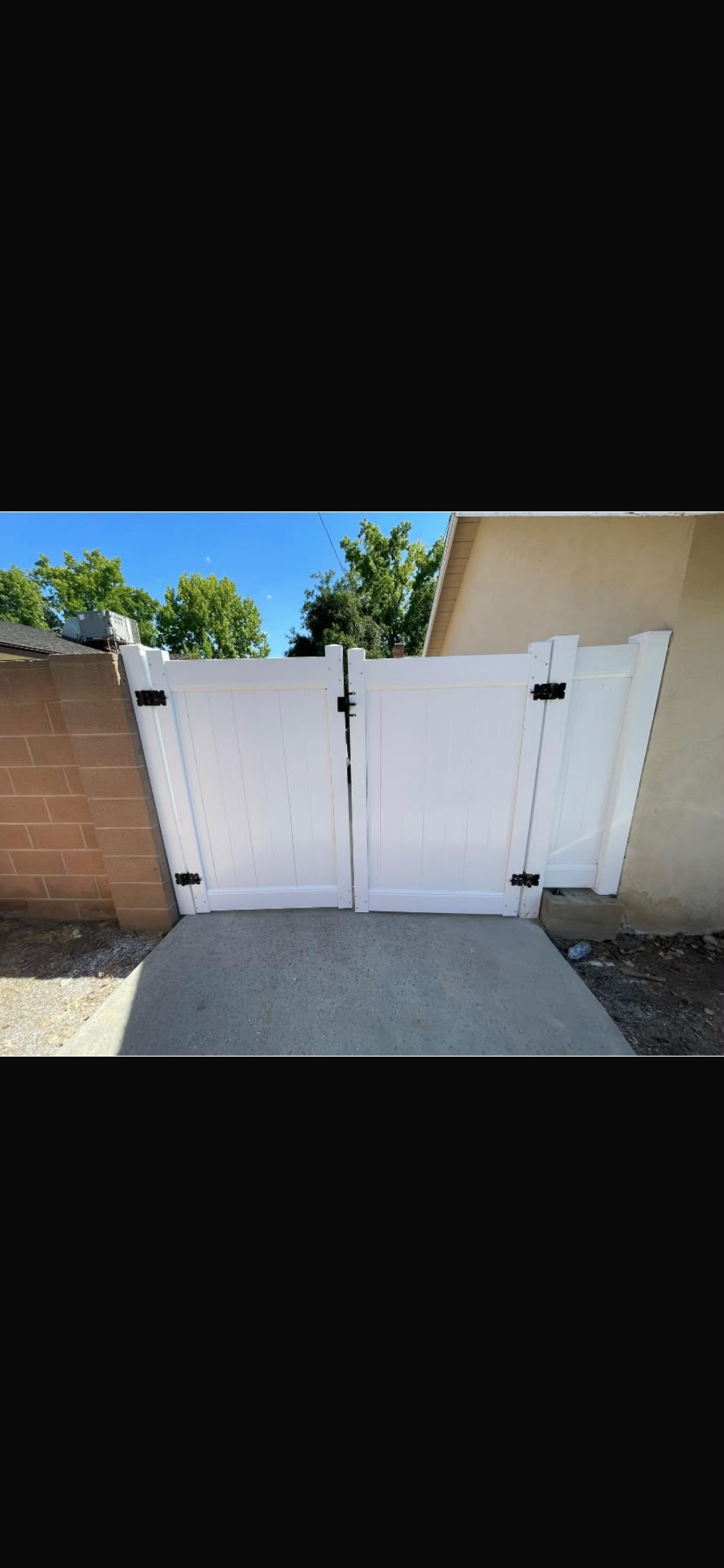 A white vinyl double gate stands open in a residential setting, flanked by a tan wall and greenery under a clear blue sky.