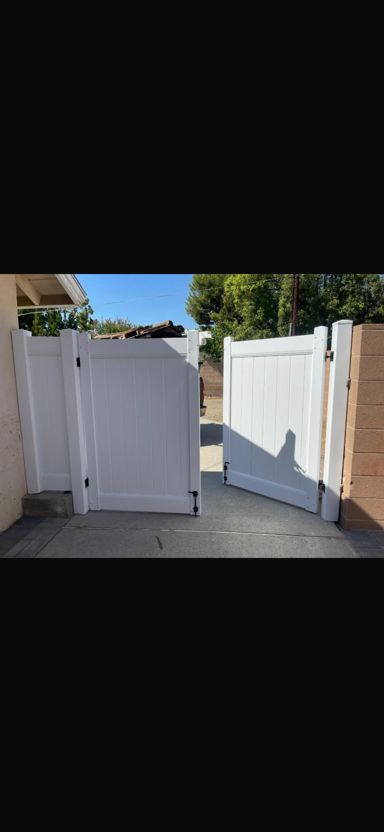 A white vinyl double gate is open, set between two fences in a sunny outdoor area with trees in the background.