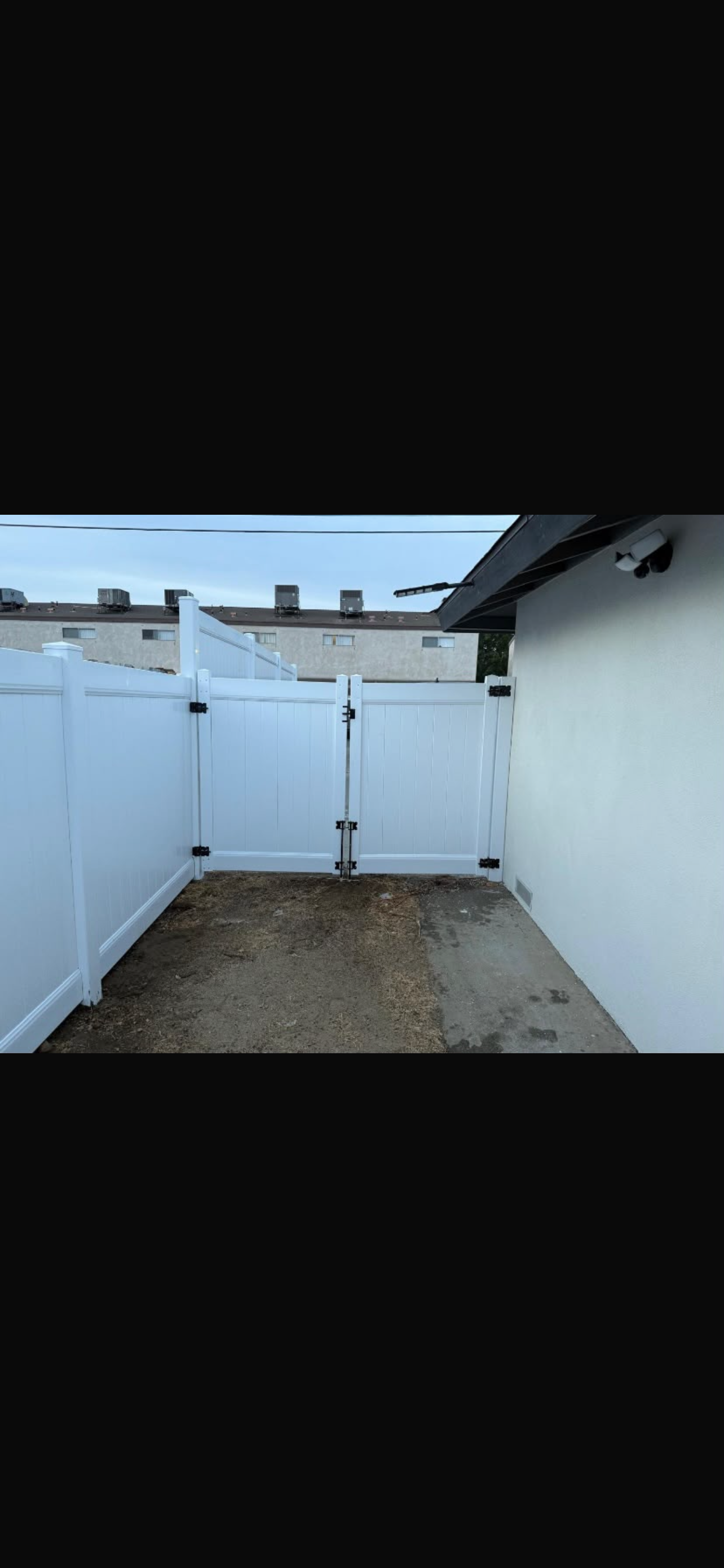 A corner view of a yard featuring white vinyl fencing and a double gate, with a gravel surface and a building wall in the background.