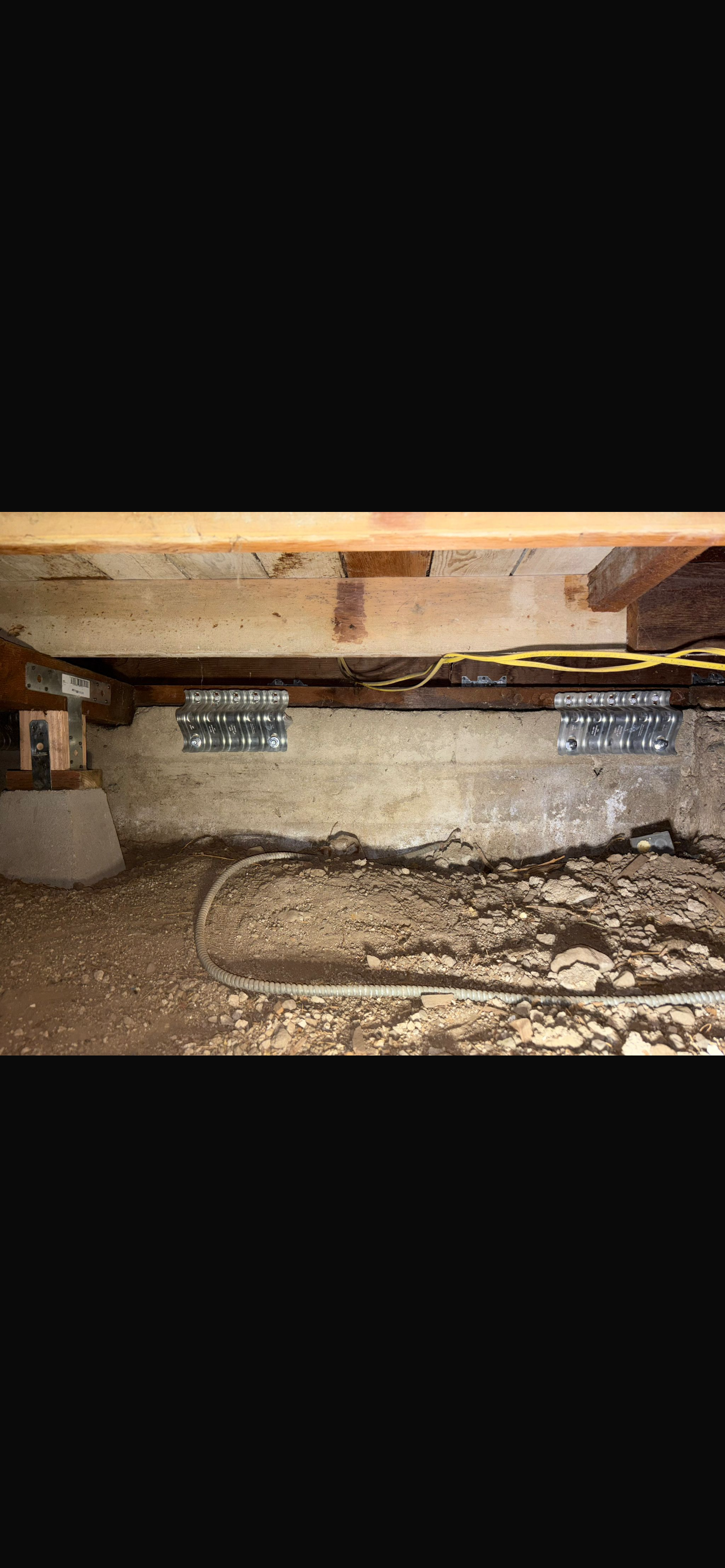 A crawl space under a house showing exposed beams, a concrete wall, and dirt flooring, with seismic retrofitting brackets visible.