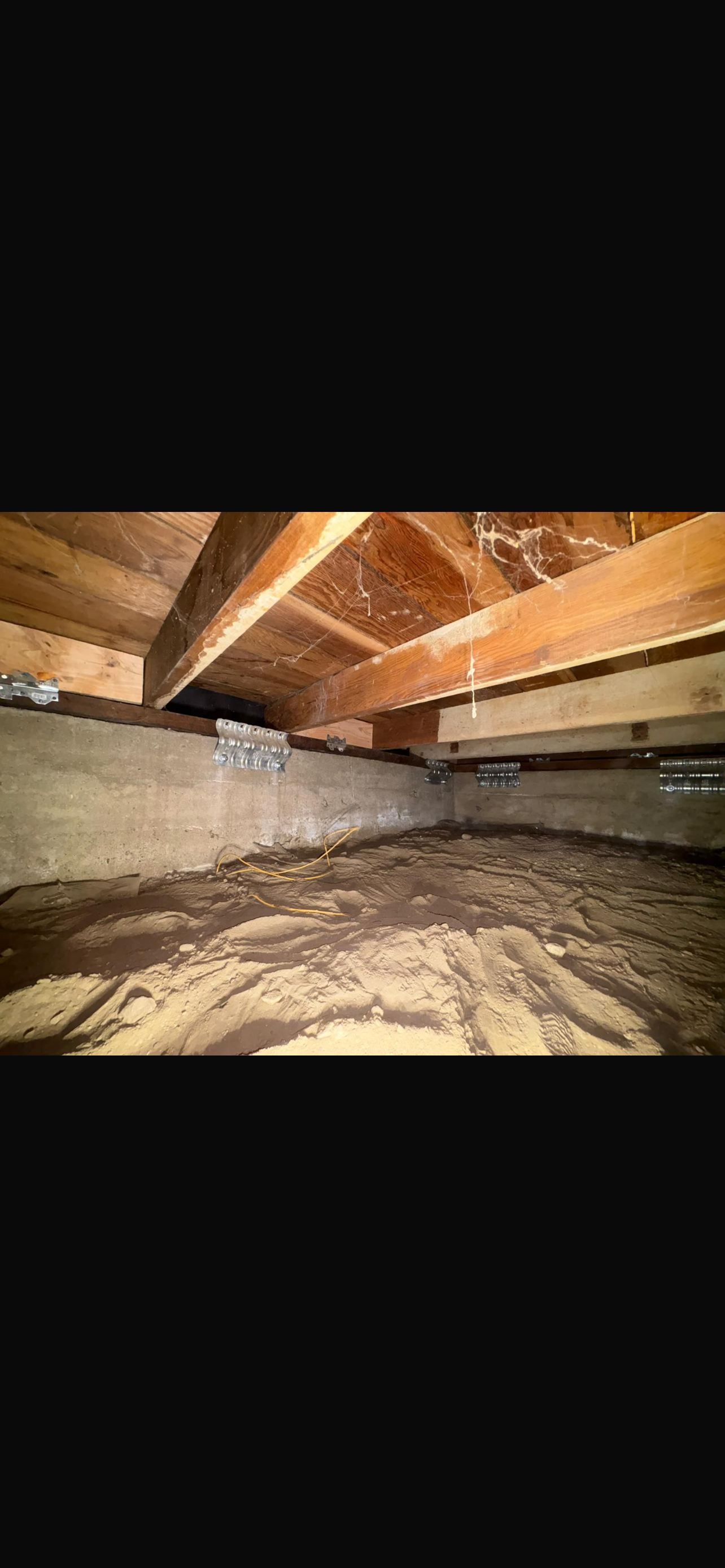 A view of a crawl space under a house, featuring wooden beams and a dirt floor, highlighting foundation maintenance concerns.