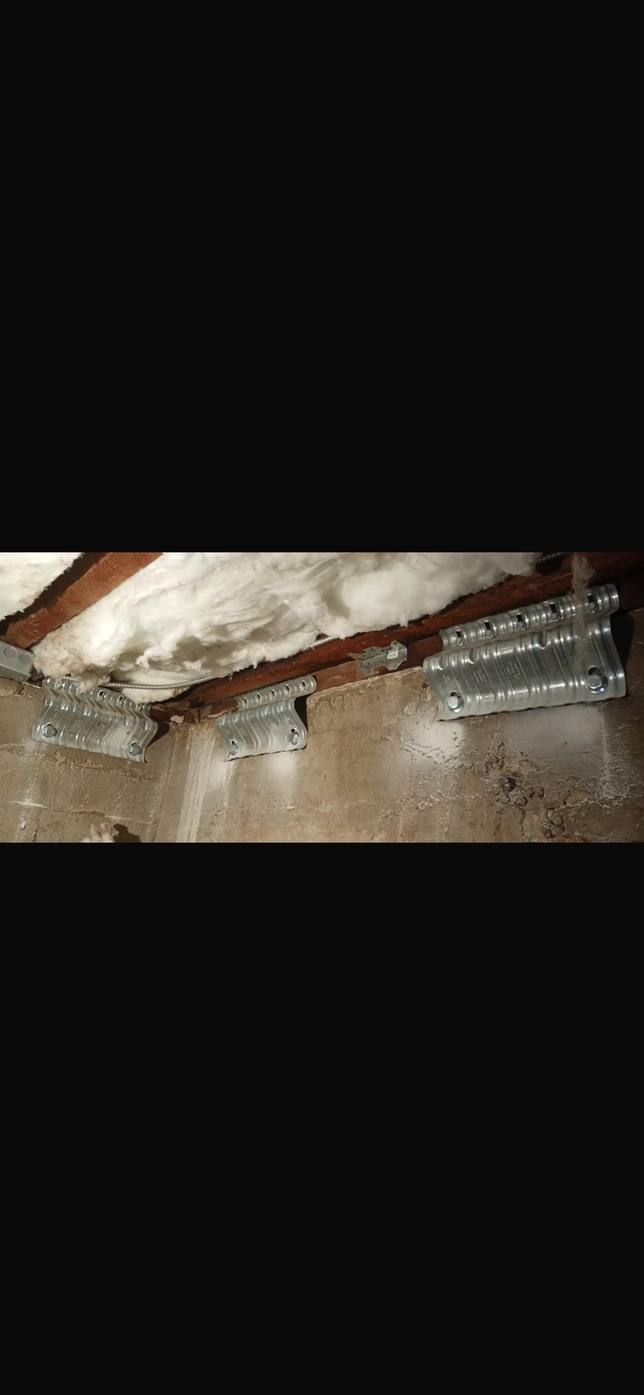 Three seismic retrofitting brackets are installed on a wooden ceiling, with insulation visible above and a textured wall below.
