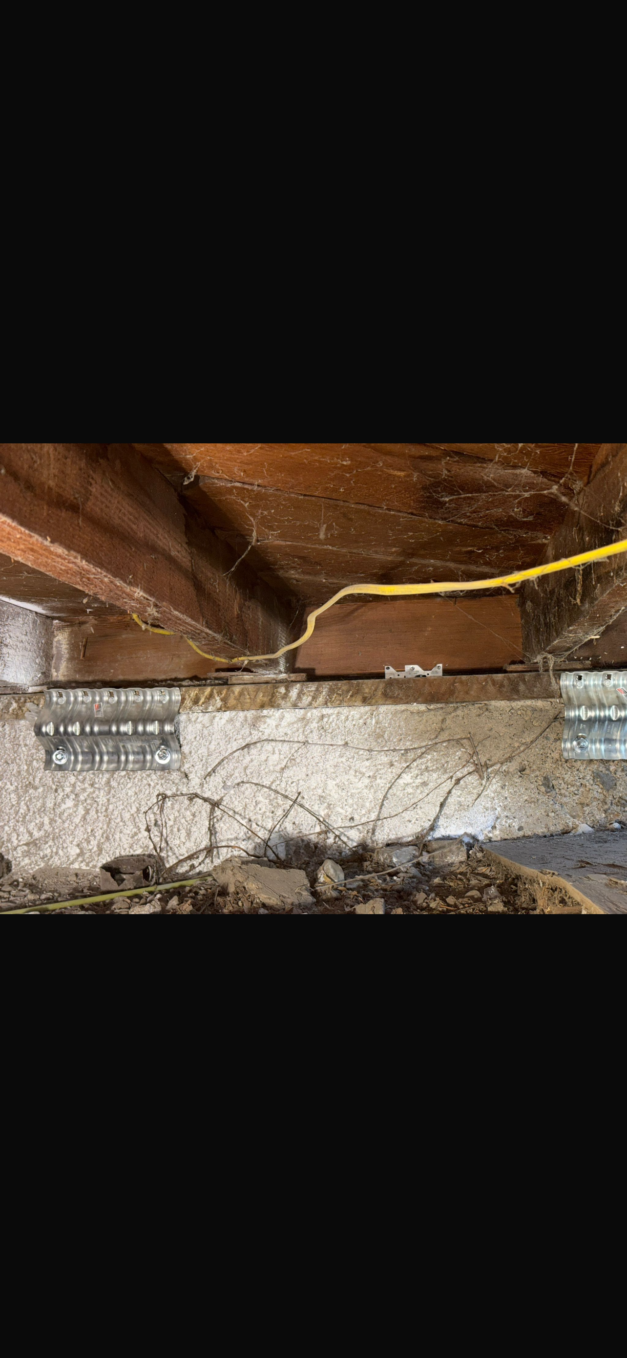 A view of a home's foundation showing exposed concrete, wooden beams, and a yellow electrical wire, with signs of wear and potential damage.