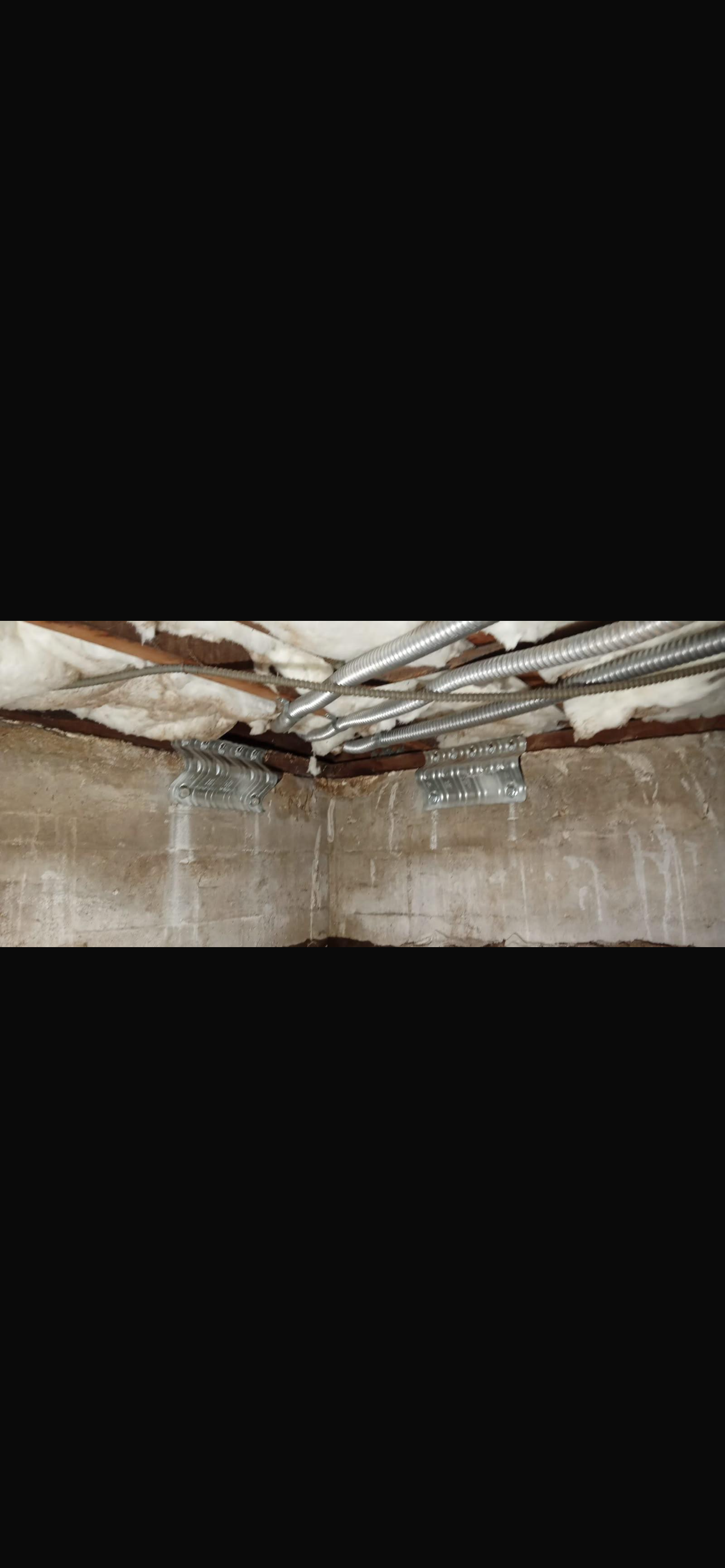 Interior view of a basement showing exposed beams, insulation, and metal pipes along the ceiling, with concrete walls.