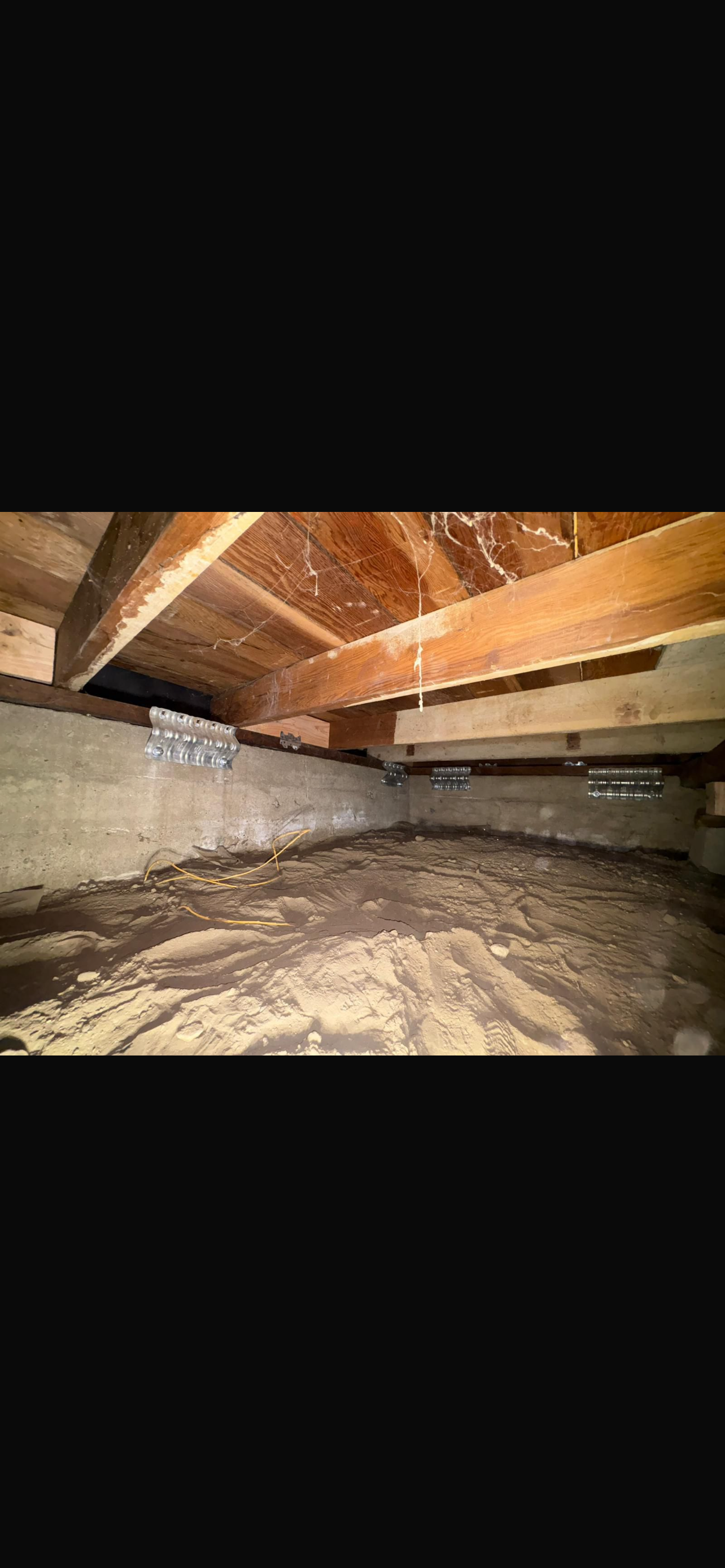 A dimly lit crawl space showing wooden beams and a concrete foundation, with dirt flooring and visible dust patterns.