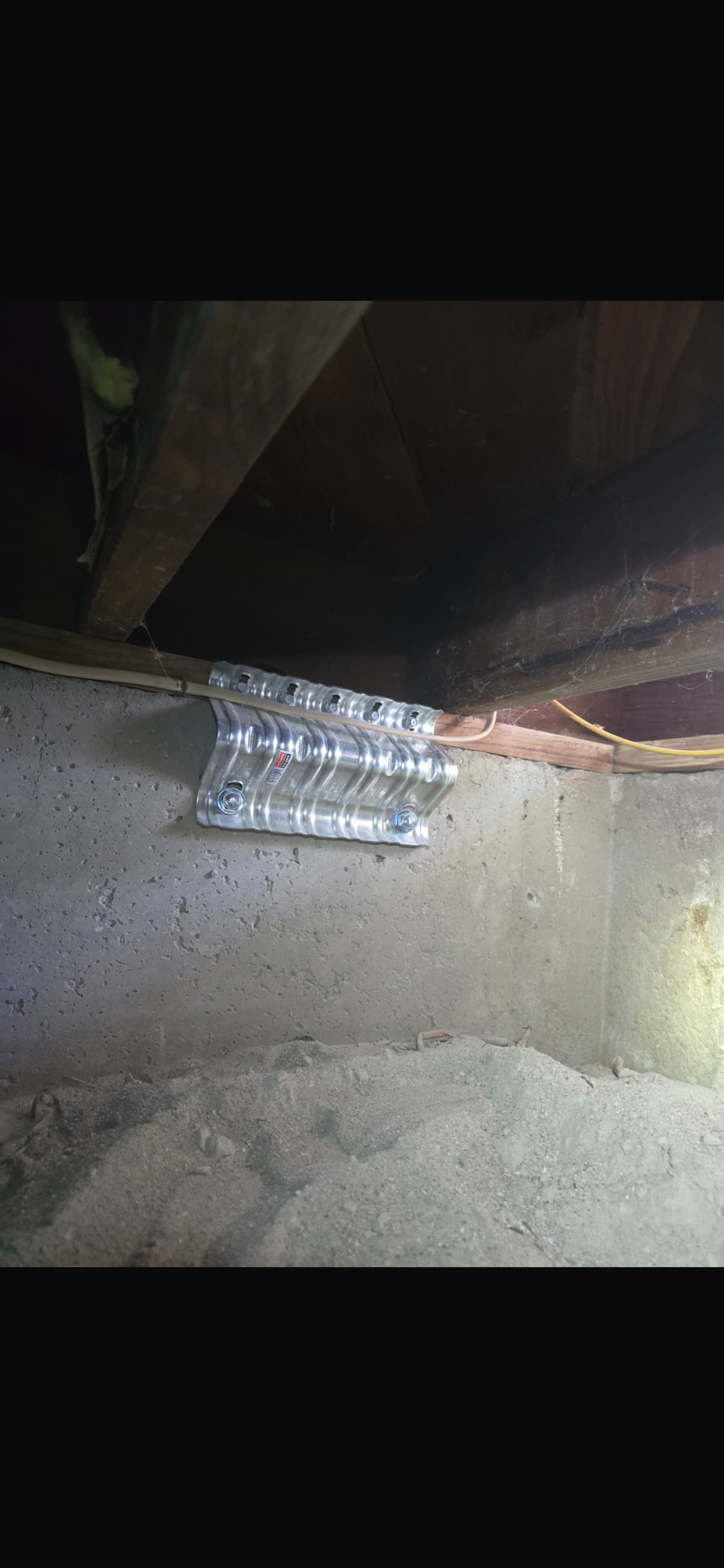 Metal seismic retrofit bracket attached to a wooden beam, with a concrete foundation and sand visible below.