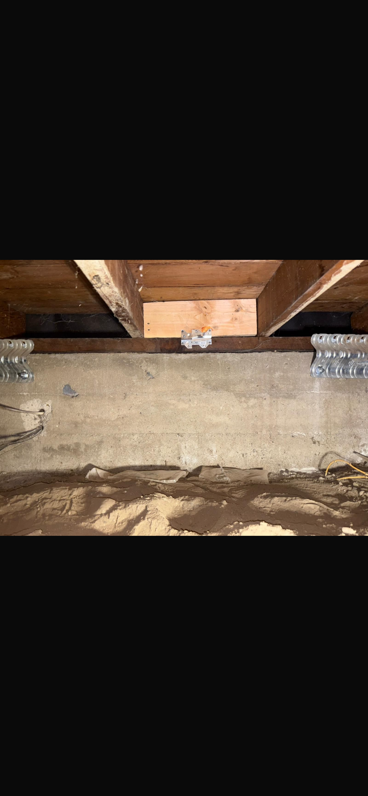 A view of a home's foundation with exposed concrete walls, wooden beams above, and dirt on the ground, highlighting structural elements.