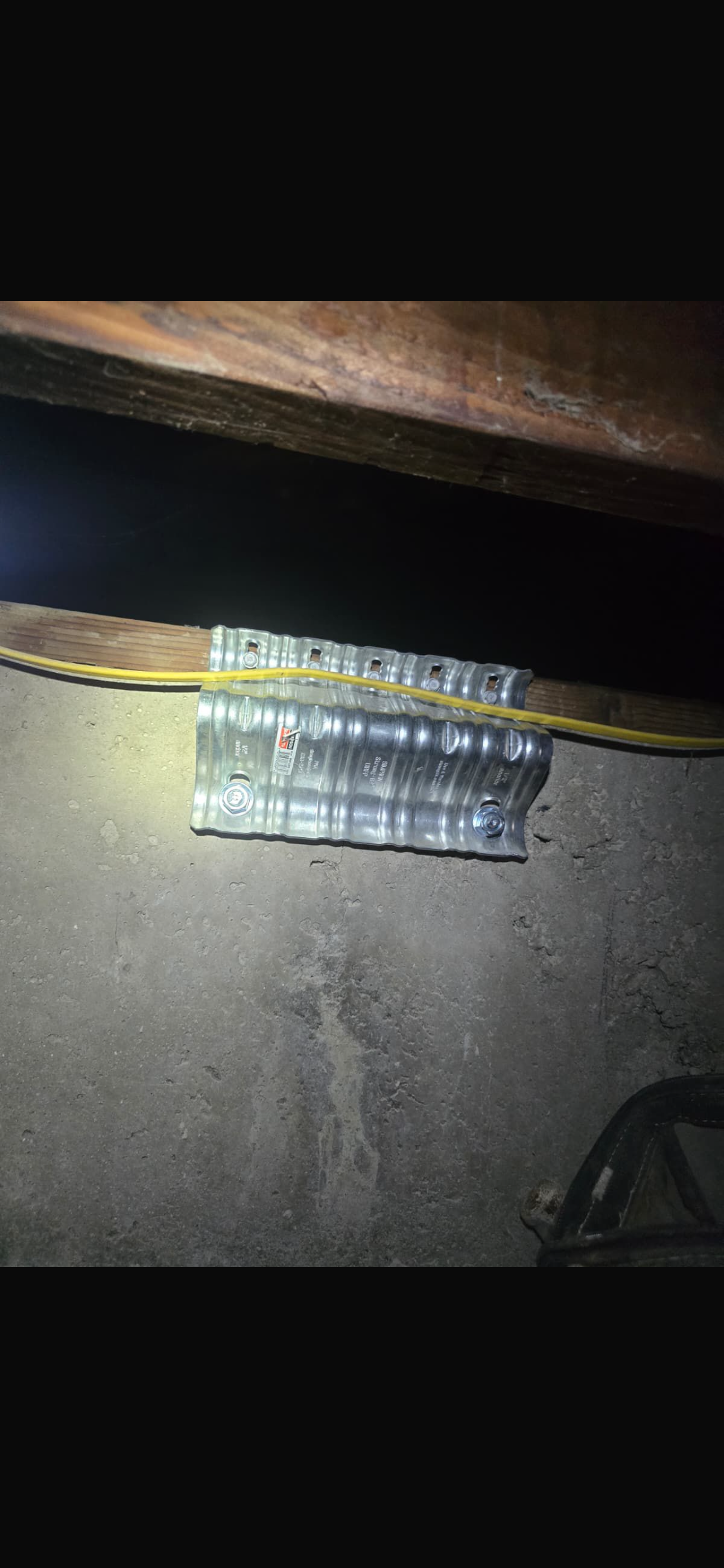 A close-up view of a foundation area showing metal brackets and wiring, with a dimly lit concrete surface beneath a wooden beam.