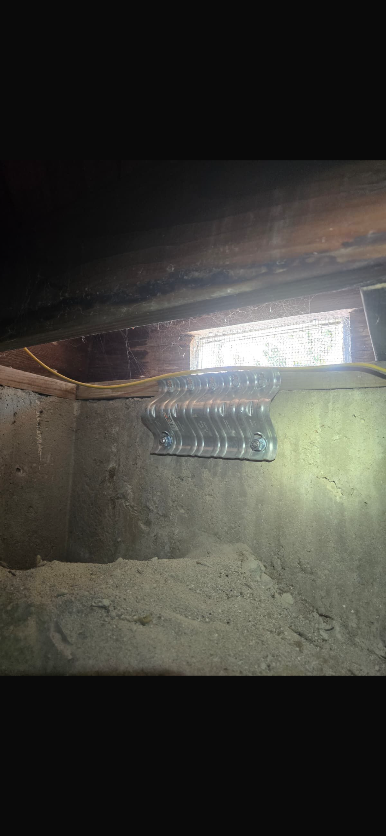 A dimly lit crawl space with exposed wooden beams, a metal bracket attached to the wall, and a small window providing light.